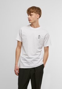 White cotton t-shirt featuring a small black graphic on the left chest. Classic crew neck with short sleeves and a straight hem.