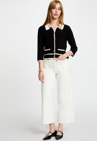Zwarte cardigan met witte rand, knoopsluiting en voorzakken, gecombineerd met witte wijde jeans en zwarte flats. Glad textuur en modern ontwerp.