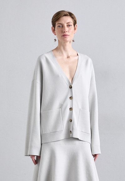 Femeie îmbrăcată cu un cardigan oversized gri deschis, cu nasturi și buzunare frontale, împreună cu o fustă asortată, pe un fundal gri simplu.
