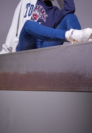 Jeans blu, felpa grigia e navy con logo, sneakers bianche, seduto su una superficie grigia con texture metallica e dettagli di ruggine.