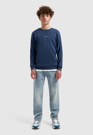 Jonge persoon met krullend haar, gekleed in een marineblauwe trui, lichtblauwe spijkerbroek en witte sneakers, staand tegen een effen achtergrond.