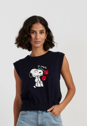 Femme aux cheveux bouclés et foncés portant un gilet sans manches bleu marine avec Snoopy et deux cerises rouges avec le texte « So sweet », associé à un jean clair.