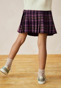 Lila-kariertes, gefaltetes Röckchen mit schwarzen und braunen Akzenten, kombiniert mit hellen Knöchelsocken und glitzernden Schuhen auf einem Holzfußboden.