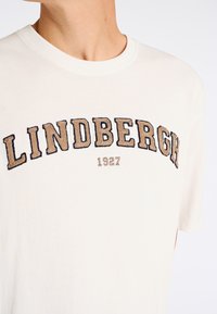 Camiseta de algodón crema con bordado texturizado "LINDBERGH" en marrón y negro sobre el año "1927". Diseño clásico de cuello redondo.