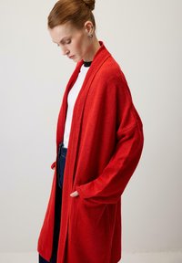 Červený nadmerný kardigán s textúrovanou tkaninou, spustenými ramenami, otvoreným predným dielom a bočnými vreckami, doplnený bielym topom a tmavými džínsami.