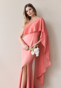 Korallefarbenes Kleid mit asymmetrischer Kapuze, seitlicher Schlitze und schlanker Silhouette; kombiniert mit weißen Blumen, die einen sanften Kontrast bilden.