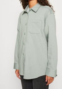 Camicia button-up verde chiaro realizzata in tessuto morbido, con colletto, taschino sul petto e maniche lunghe con polsini buttonati.