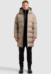 Beige pufferjakke med hætte, quiltet design og lynlås foran. Bæres over en sort top og kombineres med sorte bukser og støvler.