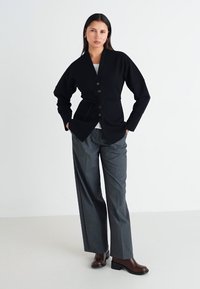 Black cardigan with a tie waist, featuring a v-neck and button closures. Worn over a gray outfit and paired with brown ankle boots.
