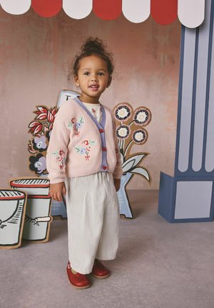 Mladá dívka v růžovém vyšívaném svetru, pruhovaných širokých kalhotách a červených botách stojí uvnitř vedle kartonových květin a pruhovaného markýzy.