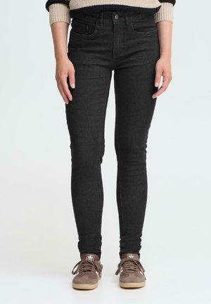 Personne portant un jean skinny noir, un pull rayé beige et noir, et des baskets Adidas marron se tenant devant un fond uni.