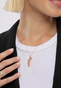 Roségouden ketting met een veerhanger en een kleine heldere steen, gedragen op een wit shirt met een accent van een zwart colbert.