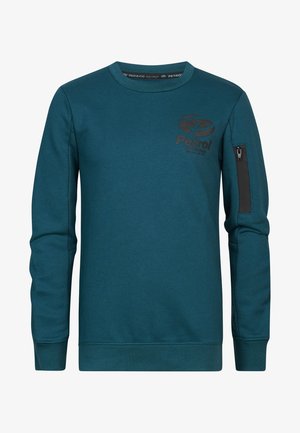 Dziecko ubrane w turkusową bluzę, szare dżinsy i białe trampki stoi na jasnym tle.