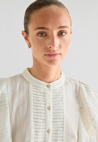 Witte blouse met een gestructureerd lijf, verticale plooien, een hoge kraag en knoopsluitingen. Zachte stof met uitlopende mouwen en een gladde afwerking.