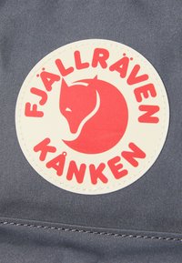 Okrogla bijela oznaka s logotipom crvene lisice i natpisom "Fjällräven Kånken" na tamnosivoj tkanini.