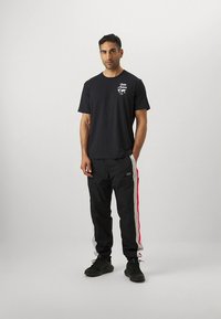 Zwart T-shirt met korte mouwen en een kleine witte afbeelding op de borst, gecombineerd met zwarte broek met rode en grijze zijstrepen. Zwarte sneakers.