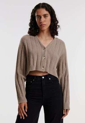 Beige Cardigan, tailliert mit gerippter Textur, V-Ausschnitt und vier Knöpfen. Kombiniert mit hoch taillierten schwarzen Jeans.