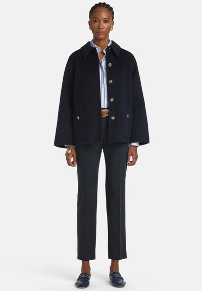Tmavomodrý kabát z vlnenej zmesi s výrazným strihom, vybavený špicatým golierom, zlatými gombíkmi a dvoma prednými vreckami. Kombinovaný s nohavicami na mieru.