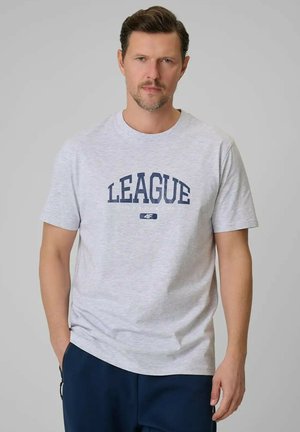 Mężczyzna z krótkimi brązowymi włosami i brodą, w jasnoszarej koszulce z nadrukiem "LEAGUE" oraz ciemnych spodniach, stojący na jednolitym szarym tle.