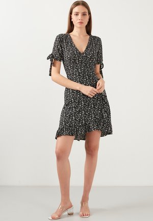 Black short dress featuring white floral print, v-neck, short sleeves with bow details, and a ruffled hem. Worn with clear heeled sandals.