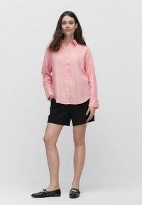 Lichtroze lange mouwen overhemd met knopen, gecombineerd met zwarte shorts en zwarte ballerina's. Het overhemd heeft een klassieke kraag en een ontspannen pasvorm.