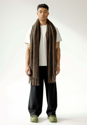 Brown wool scarf with fringed ends, worn over a light t-shirt, paired with wide black ribbed pants and olive green sneakers.