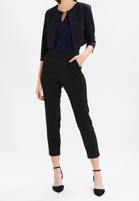 Schwarze, taillierte Blazerjacke über einem marineblauen Oberteil, kombiniert mit hochtaillierten, schwarzen, schmal geschnittenen Hosen und schwarzen, spitz zulaufenden Schuhen mit Knöchelriemen.