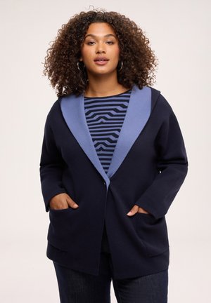 Woman with curly hair wearing a navy coat with light blue collar over a black and blue striped shirt, hands in coat pockets.