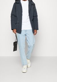 Donkerblauwe geïsoleerde jas met een capuchon, ritssluiting aan de voorkant en zakken, gecombineerd met lichtblauwe jeans en witte sneakers, terwijl een zwarte tas wordt vastgehouden.