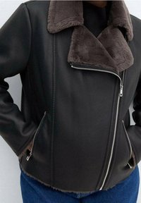 Veste en cuir noire avec un col en fausse fourrure marron, fermeture éclair asymétrique et poches latérales. Matériau texturé et design ajusté.