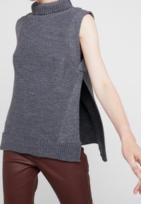 Maglione grigio lavorato a maglia con collo alto, design senza maniche, spacchi laterali e orlo a coste. Tessuto morbido e vestibilità ampia, abbinato a pantaloni marroni.