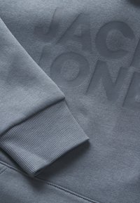 Harmaa huppari, jossa on teksturoitu kangas, ribattu hihansuu ja suuri painettu teksti "JACK & JONES" matchaavassa sävyssä.