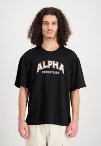 Fekete, túlméretezett pamut póló. Nagy fehér és narancssárga "ALPHA" felirattal rendelkezik, alatta "INDUSTRIES" felirattal. Rövid ujjú, kerek nyakkivágású.