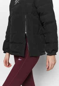 Personne portant une doudoune noire avec la fermeture éclair partiellement ouverte, assortie à des leggings bordeaux avec un petit logo blanc sur la cuisse.
