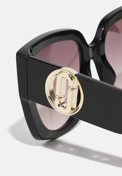 Schwarze übergroße Sonnenbrille mit Verlaufsgläsern, die ein auffälliges goldenes, rundes Logo auf dem Rahmen aufweist. Glatte, glänzende Oberfläche.