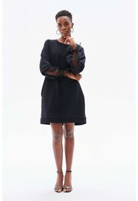 Schwarzes Kleid mit lockerer Passform, das durchsichtige, puffige Ärmel mit kleinen Perlen aufweist. Getragen mit Riemchensandalen, die eine glatte Textur zeigen.