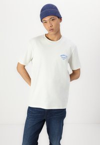 Fehér pamut póló kék "Wrangler Jeans 1947" felirattal a bal mellrészen, sötétkék farmernadrággal és csíkos kék sapkával párosítva.