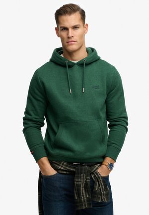 Man met kort haar draagt een groene hoodie en blauwe spijkerbroek, met een geruit overhemd gestrikt om zijn middel en een horloge aan zijn linker pols.