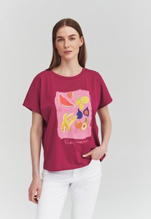 Frau trägt ein magentafarbenes T-Shirt mit bunten Fruchtillustrationen und dem Text "Fruity summer", kombiniert mit weißen Hosen, stehend vor weißem Hintergrund.