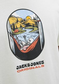 Balta medvilninė marškinėliai su spalvinga grafika, vaizduojančia kaukolę hamake, aplink kurią matyti kalnai, upė ir saulėlydis.