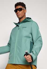 Chaqueta verde menta resistente al agua con capucha, cierre frontal con cremallera, detalles en negro, puños elásticos y logo en el pecho. Sostenida con un bastón de senderismo.