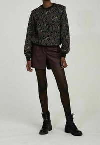 Czarna bluza w wzory z kolorowym motywem paisley, zestawiona z ciemnoczerwonymi spodniami szortami z ekologicznej skóry, czarnymi rajstopami w kropki oraz czarnymi sznurowanymi botkami.