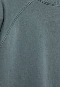 Gråblå sweatshirt med struktureret stof, ribbet halsudskæring og synlige søm; sammensætningen fremstår blød og afslappet.