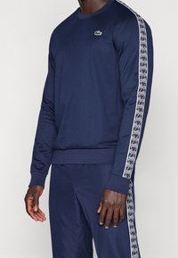 Námořnická mikina s dlouhými rukávy, žebrovanými manžetami a spodním lemem. Obsahuje bílý geometrický vzor podél rukávů a malé zelené logo na hrudi.