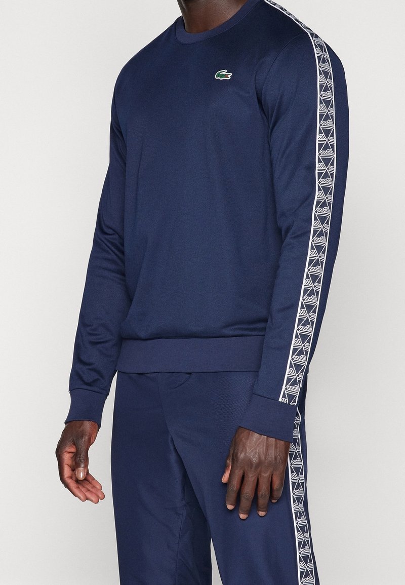 Námořnická mikina s dlouhými rukávy, žebrovanými manžetami a spodním lemem. Obsahuje bílý geometrický vzor podél rukávů a malé zelené logo na hrudi.