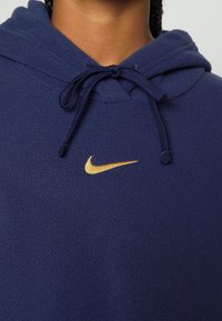 Mörkblå fleecehoodie med dragsko i huvan, strukturerad väv och en gyllene Nike-logga på bröstet. Enkel och funktionell design.