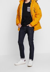 Giacca isolante gialla, maglione nero con tasca a motivo, jeans scuri e sneaker bianche. Tessuto strutturato, design aderente.