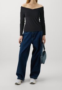 Black off-the-shoulder long-sleeve top paired with loose blue denim jeans and white sneakers, holding a small denim handbag.