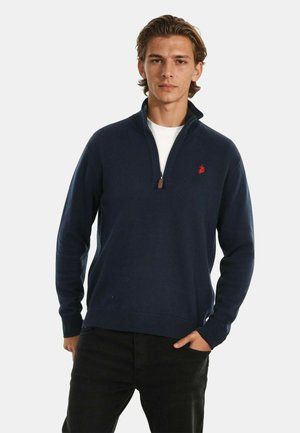 Junger Mann in einem navyblauen Quarter-Zip-Pullover mit rotem Logo, weißem Unterhemd und schwarzen Hosen, der mit einer Hand in der Tasche steht.