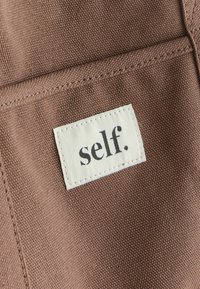 Brązowy materiał o teksturowanym splocie, z kremową metką, na której widnieje wyraz "self." napisany tłustym czarnym fontem. Widoczne są wystające krawędzie.